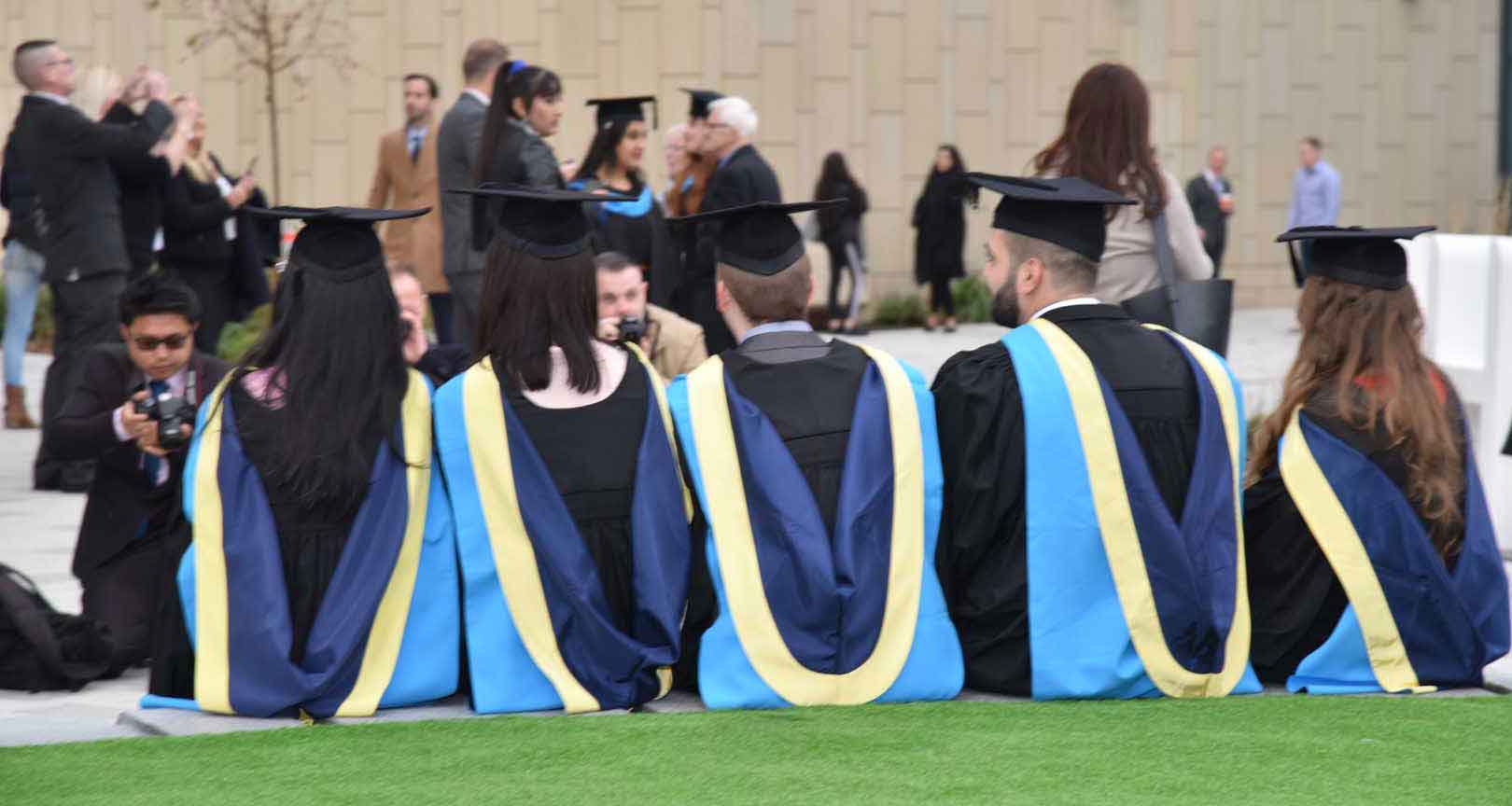 the back of 4 graduates wearing their cap and gowns, sat down in the plaza